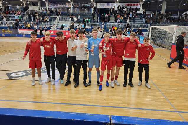 Gruppe von zehn jungen Fu&szlig;ballspielern in roten Trikots und Torwart in Blau posieren nach einem Spiel in einer Halle.