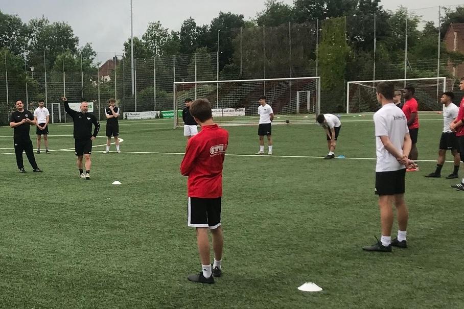 Gruppentraining auf einem Fu&szlig;ballplatz, Trainer steht vor einer Versammlung von Spielern in unterschiedlichen Trikots.