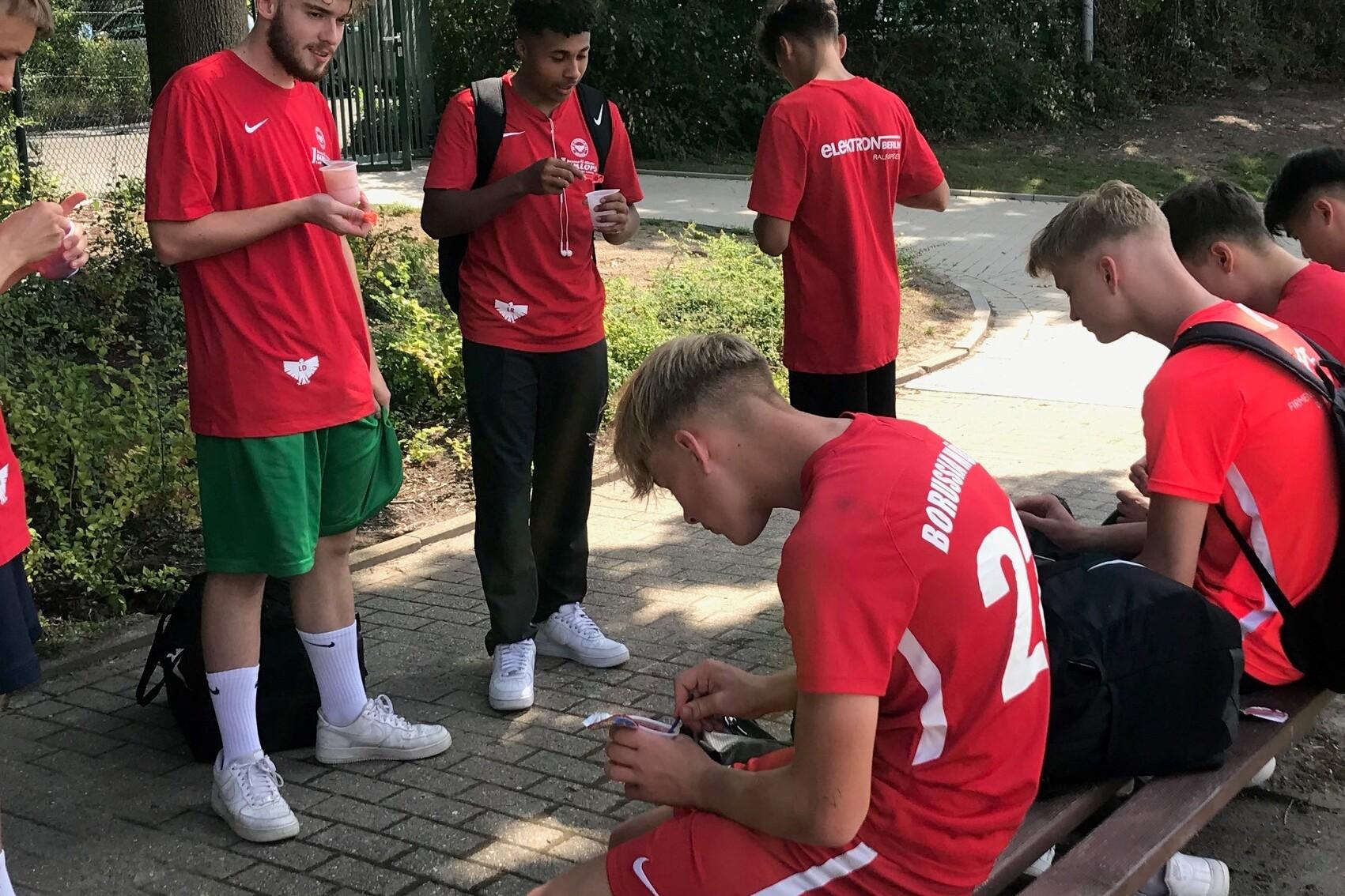 Gruppe junger Sportler in roten Trikots sitzt auf einer Bank und snackt, umgeben von B&auml;umen und einem Weg.