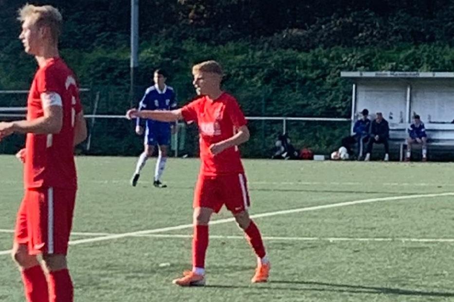 Zwei Fu&szlig;ballspieler in roten Trikots stehen auf einem Rasenplatz, w&auml;hrend im Hintergrund weitere Spieler sitzen.