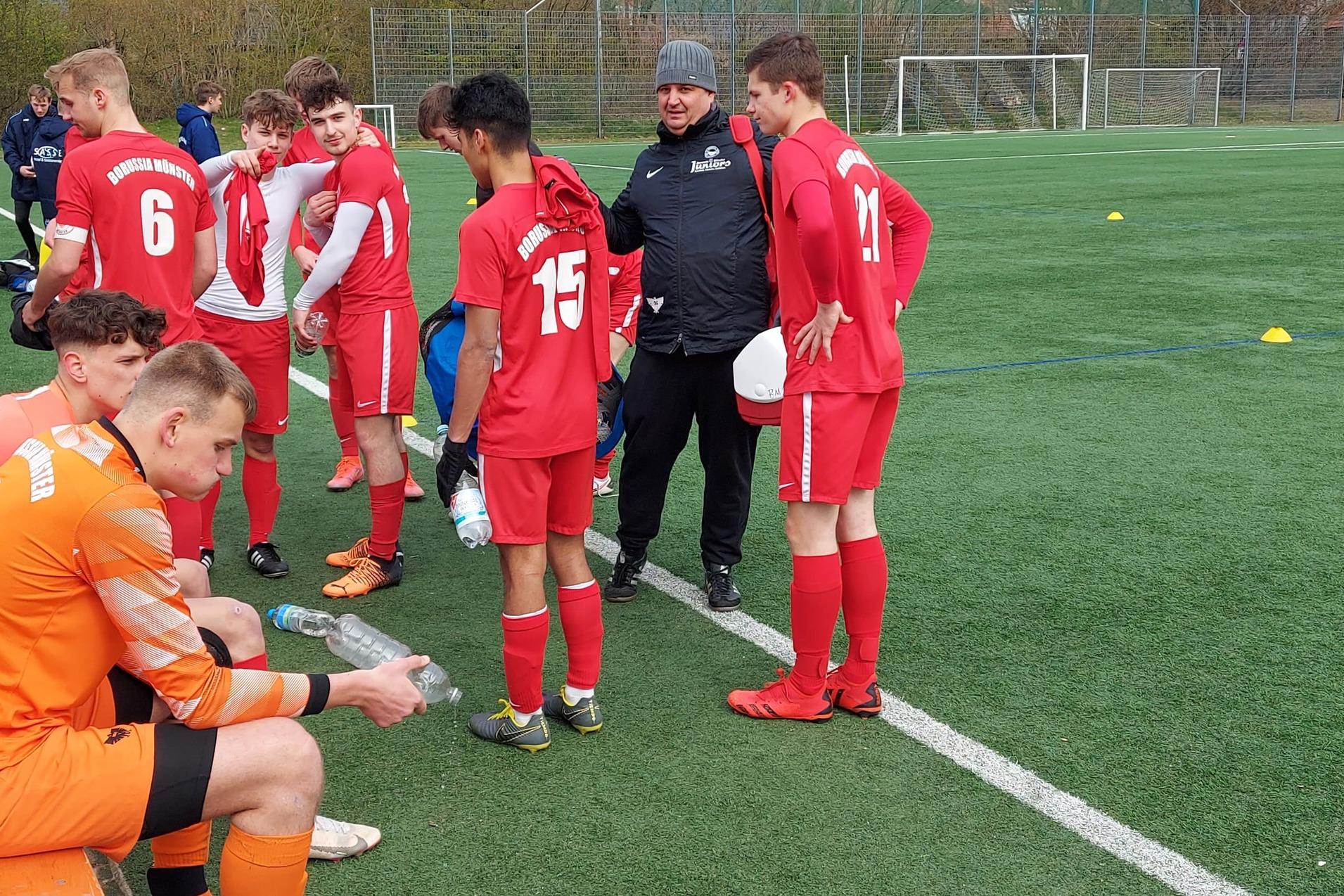 Fu&szlig;ballmannschaft in roten Trikots versammelt sich am Spielfeldrand. Ein Trainer spricht mit den Spielern.