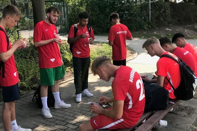 Gruppe junger Sportler in roten Trikots sitzt auf einer Bank und snackt, umgeben von B&auml;umen und einem Weg.
