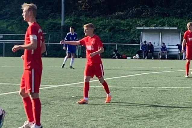 Zwei Fu&szlig;ballspieler in roten Trikots stehen auf einem Rasenplatz, w&auml;hrend im Hintergrund weitere Spieler sitzen.