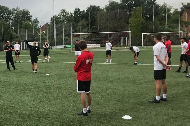 Gruppentraining auf einem Fu&szlig;ballplatz, Trainer steht vor einer Versammlung von Spielern in unterschiedlichen Trikots.