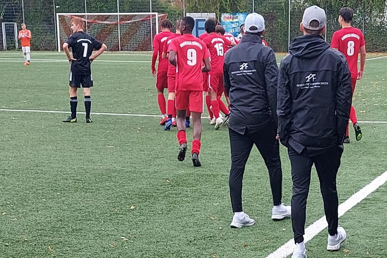 Fu&szlig;ballspieler in roten Trikots betreten das Spielfeld, w&auml;hrend Trainer und Schiedsrichter am Rand stehen.