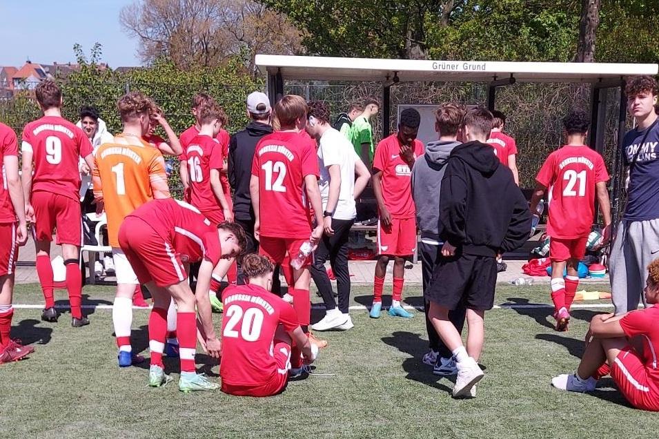 Junge Fu&szlig;ballspieler mit roten Trikots stehen und sitzen auf einem Sportplatz, eine Bank im Hintergrund.