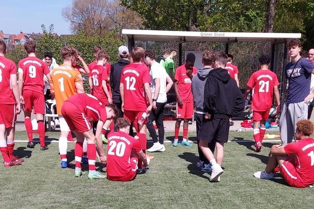 Junge Fu&szlig;ballspieler mit roten Trikots stehen und sitzen auf einem Sportplatz, eine Bank im Hintergrund.