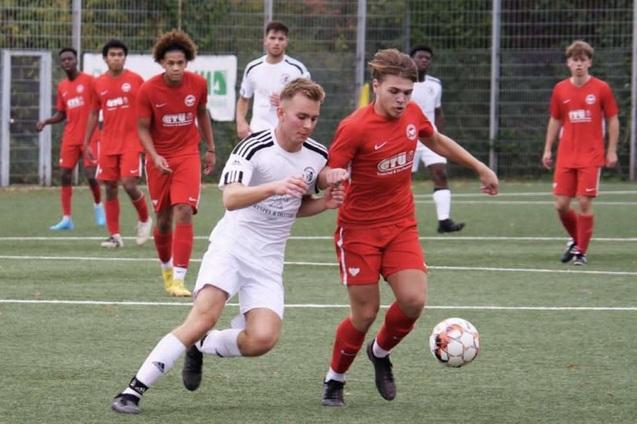 Zwei Fu&szlig;ballspieler k&auml;mpfen um den Ball auf einem Kunstrasenplatz, w&auml;hrend mehrere Spieler im Hintergrund zuschauen.