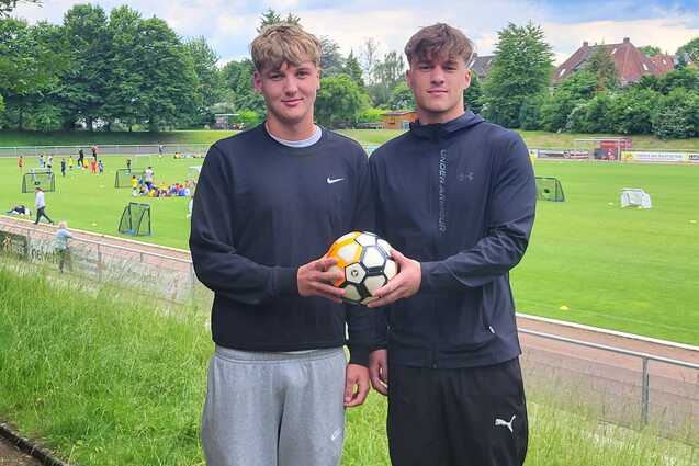 Zwei junge M&auml;nner halten einen Fu&szlig;ball, im Hintergrund ein Trainingsgel&auml;nde mit weiteren Spielern und Toren.
