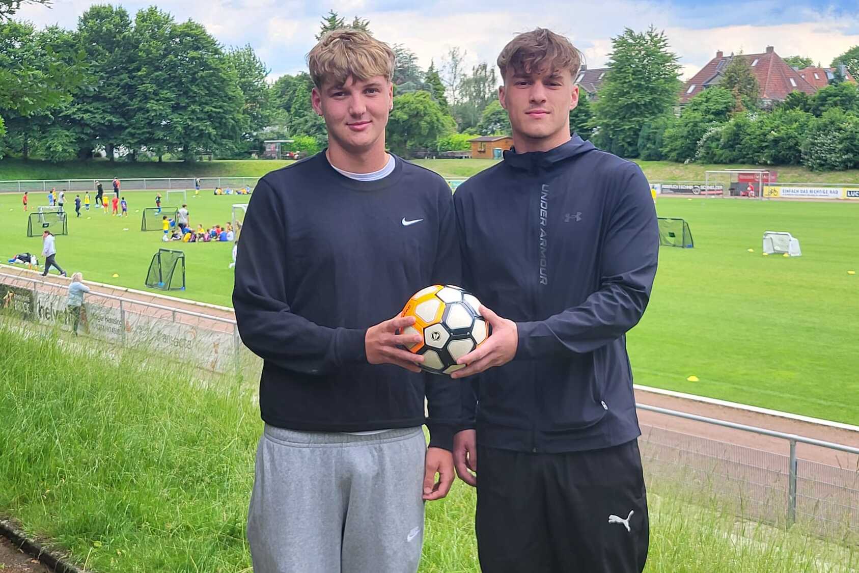 Zwei junge M&auml;nner halten einen Fu&szlig;ball, im Hintergrund ein Trainingsgel&auml;nde mit weiteren Spielern und Toren.