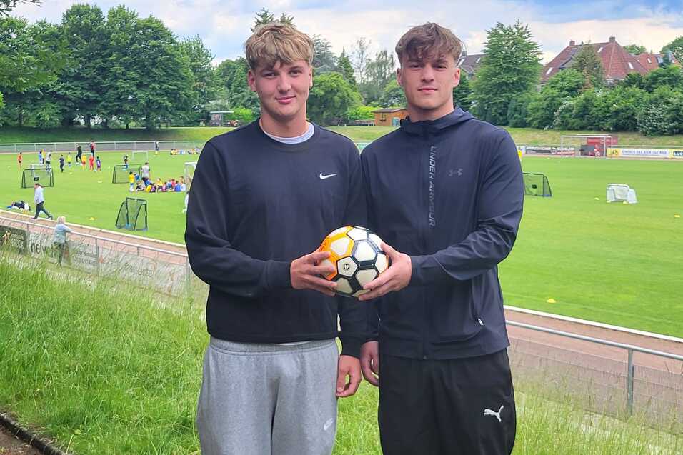 Zwei junge M&auml;nner halten einen Fu&szlig;ball, im Hintergrund ein Trainingsgel&auml;nde mit weiteren Spielern und Toren.