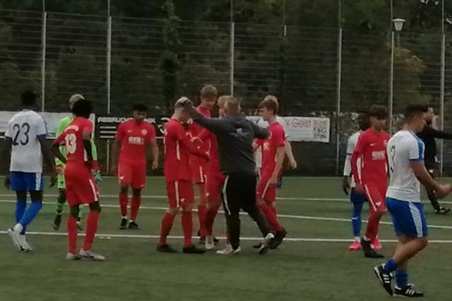 Gruppierung von Fu&szlig;ballspielern in roten Trikots auf dem Feld, w&auml;hrend ein Schiedsrichter die Situation beobachtet.