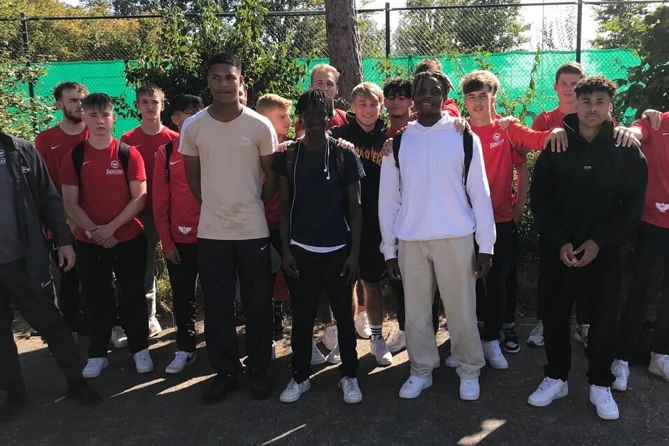 Gruppe von Jugendlichen in Sportkleidung steht vor einem Tennisplatz mit gr&uuml;ner Umz&auml;unung.