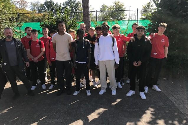 Gruppe von Jugendlichen in Sportkleidung steht vor einem Tennisplatz mit gr&uuml;ner Umz&auml;unung.