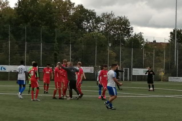 Gruppierung von Fu&szlig;ballspielern in roten Trikots auf dem Feld, w&auml;hrend ein Schiedsrichter die Situation beobachtet.