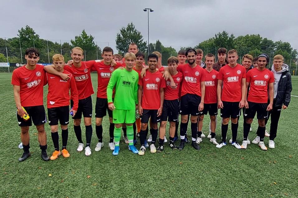 Gruppe von 15 Fu&szlig;ballspielern in roten Trikots steht auf einem Kunstrasenplatz, umgeben von B&auml;umen und einem Sportplatz.