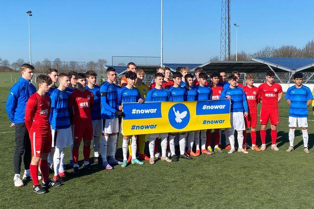 Gruppenbild von Fu&szlig;ballspielern in blauen und roten Trikots, die ein Banner mit der Aufschrift "#nowar" halten.