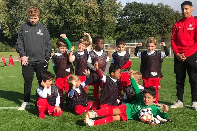 Gruppe von Kindern in Fu&szlig;balltrikots posiert l&auml;chelnd auf einem gr&uuml;nen Sportplatz, zwei Trainer stehen hinter ihnen.