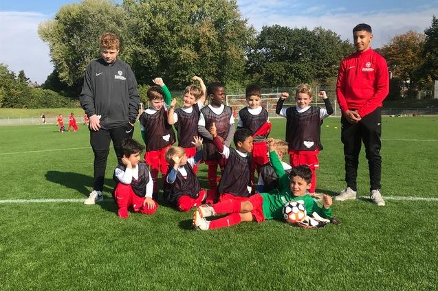 Gruppe von Kindern in Fu&szlig;balltrikots posiert l&auml;chelnd auf einem gr&uuml;nen Sportplatz, zwei Trainer stehen hinter ihnen.