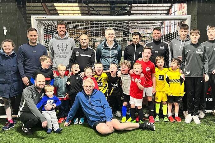 Gruppenfoto von Trainern und Kindern in Sportkleidung auf einem Fu&szlig;ballplatz, umgeben von einem Tor und Netz.