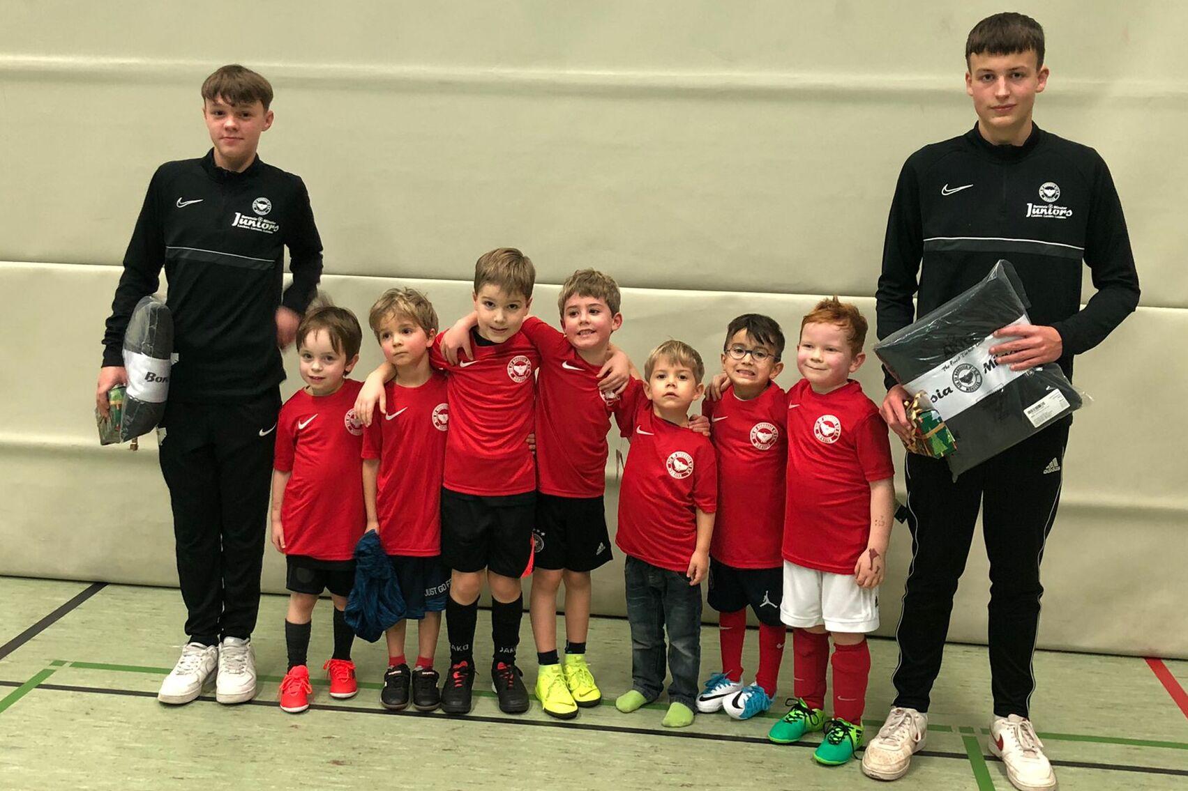Gruppe von zehn Kindern in roten Trikots, zwei Trainer in Schwarz, stehen in einer Sporthalle mit neutralem Hintergrund.