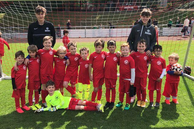 Gruppe von 14 Kindern in roten Fu&szlig;balltrikots und zwei Trainern vor einem Tor auf einem Fu&szlig;ballfeld.