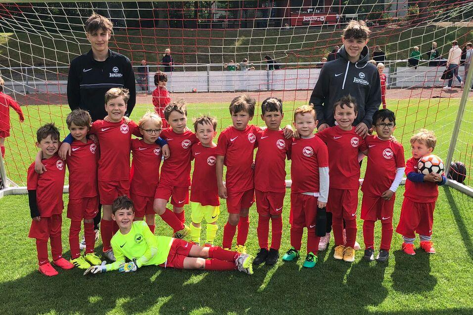 Gruppe von 14 Kindern in roten Fu&szlig;balltrikots und zwei Trainern vor einem Tor auf einem Fu&szlig;ballfeld.