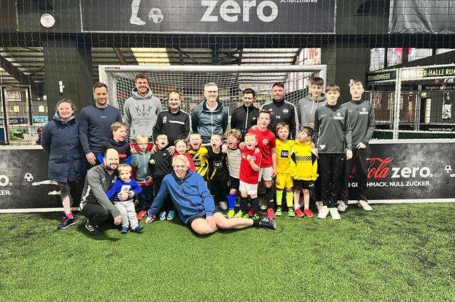 Gruppenfoto von Trainern und Kindern in Sportkleidung auf einem Fu&szlig;ballplatz, umgeben von einem Tor und Netz.