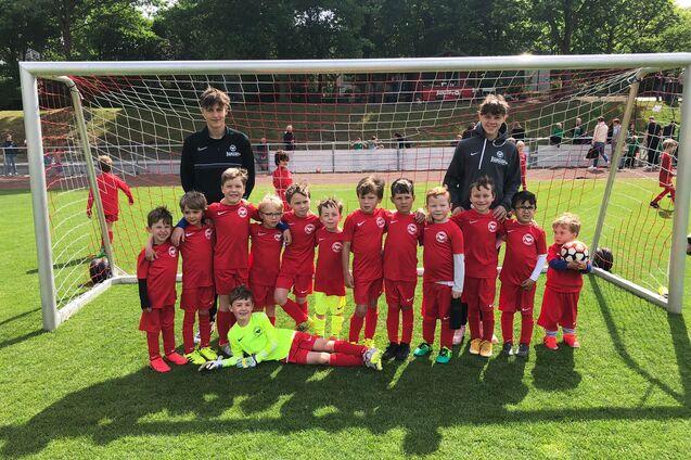 Gruppe von 14 Kindern in roten Fu&szlig;balltrikots und zwei Trainern vor einem Tor auf einem Fu&szlig;ballfeld.