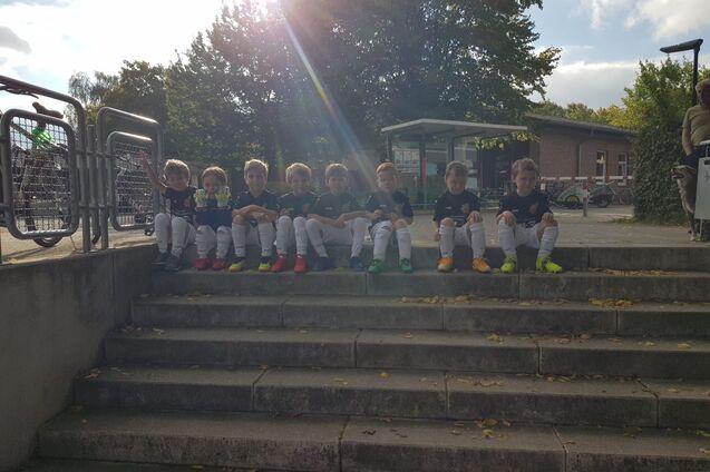 Gruppe von Kindern im Sportoutfit sitzt auf Treppenstufen in einem Au&szlig;enbereich, mit B&auml;umen im Hintergrund.