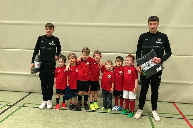 Gruppe von zehn Kindern in roten Trikots, zwei Trainer in Schwarz, stehen in einer Sporthalle mit neutralem Hintergrund.