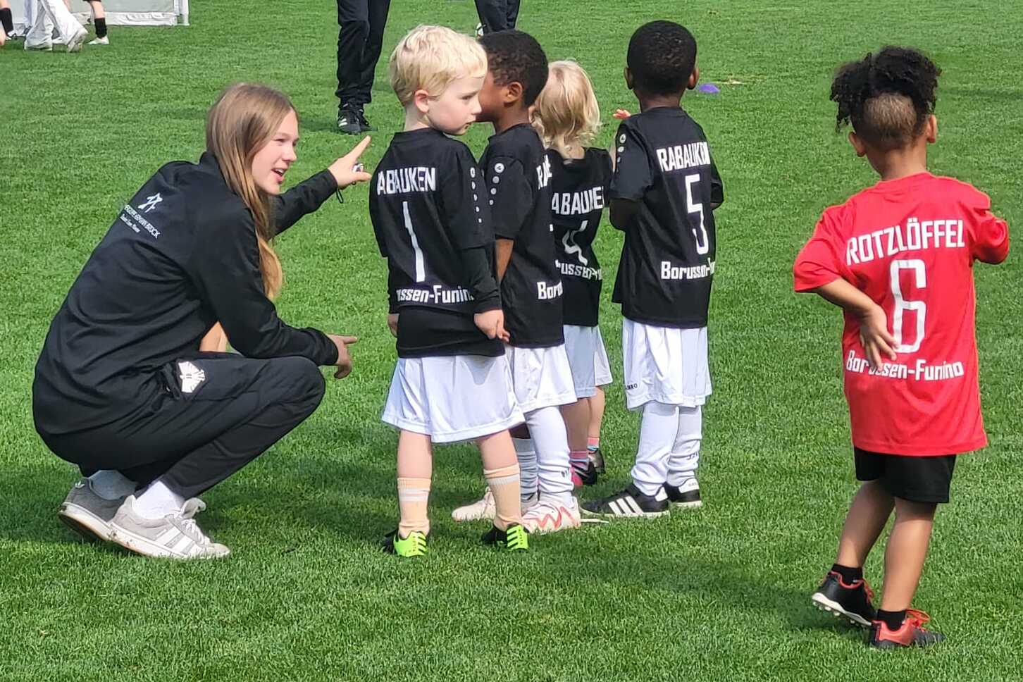 Trainerin spricht mit einer Gruppe von Kindern in Fu&szlig;balltrikots auf einem Sportplatz.
