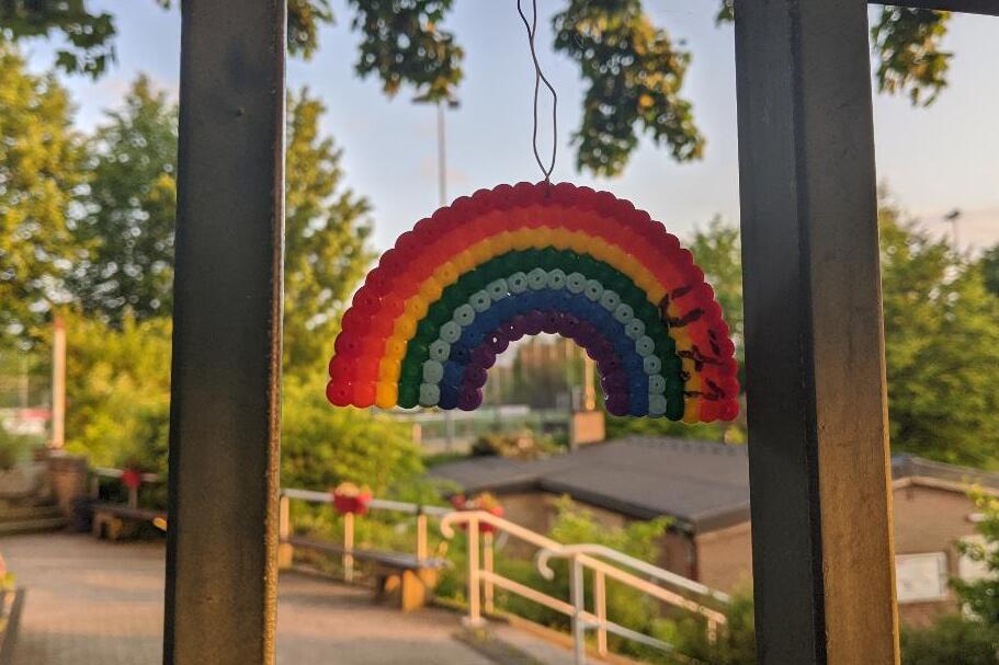 Bunter Regenbogen aus Perlen h&auml;ngt an einem Metallgel&auml;nder, im Hintergrund B&auml;ume und ein Weg zu einem Geb&auml;ude.