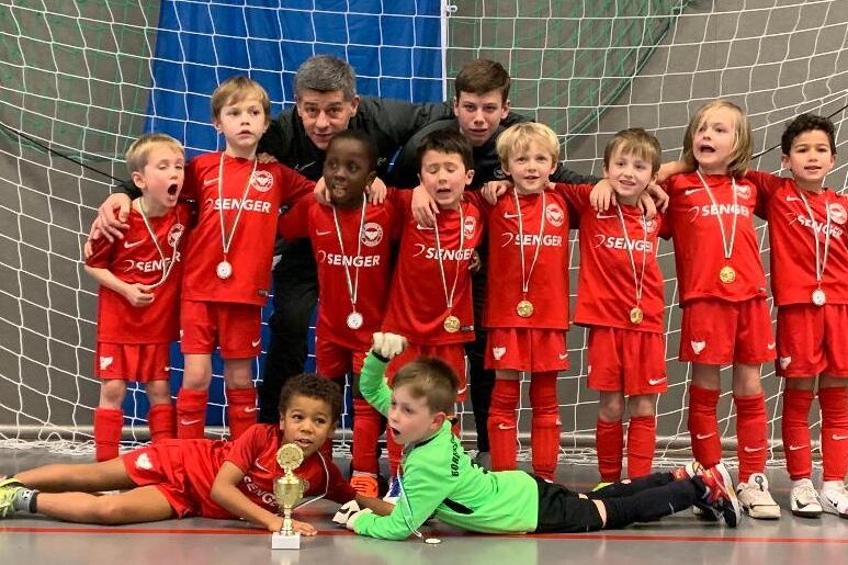 Eine Gruppenaufnahme von zw&ouml;lf jubelnden Kindern in roten Fu&szlig;balltrikots und zwei Trainern vor einem Tor, mit Medaillen.