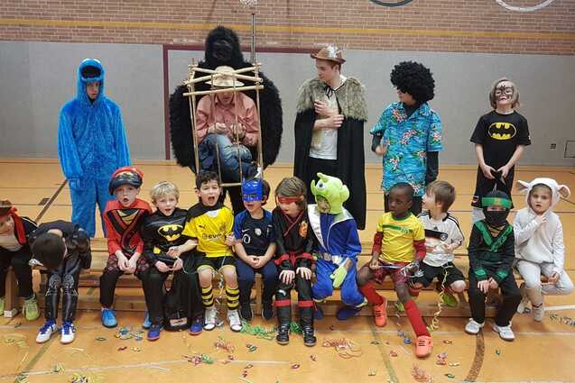 Gruppe von Kindern in Kost&uuml;men, die in einer Halle auf einer Bank sitzen, mit Konfetti auf dem Boden.