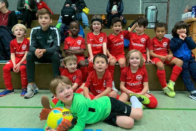 Gruppe von Kindern in roten Fu&szlig;balltrikots, sitzend auf einer Trib&uuml;ne, l&auml;cheln in die Kamera. Ein Junge tr&auml;gt ein Torwart-Trikot.