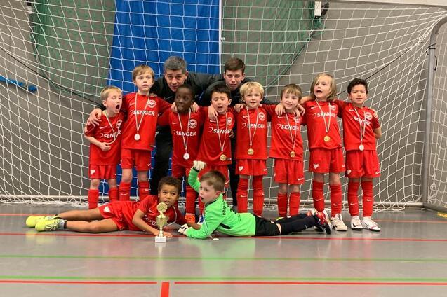Eine Gruppenaufnahme von zw&ouml;lf jubelnden Kindern in roten Fu&szlig;balltrikots und zwei Trainern vor einem Tor, mit Medaillen.