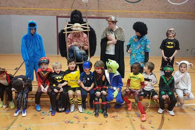 Gruppe von Kindern in Kost&uuml;men, die in einer Halle auf einer Bank sitzen, mit Konfetti auf dem Boden.