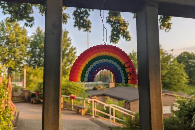 Bunter Regenbogen aus Perlen h&auml;ngt an einem Metallgel&auml;nder, im Hintergrund B&auml;ume und ein Weg zu einem Geb&auml;ude.