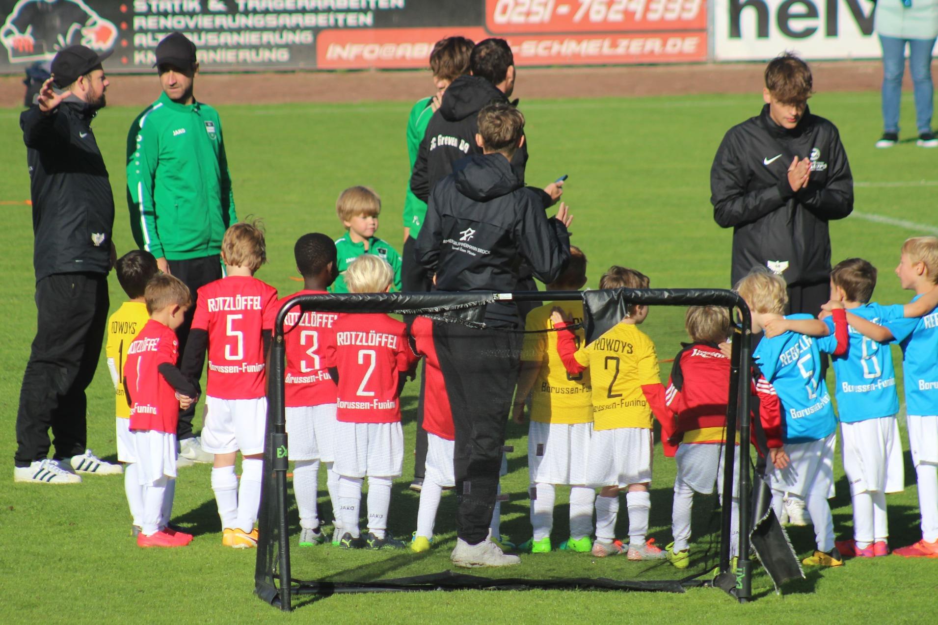 Zwei Gruppen von Kindern in Fu&szlig;balltrikots stehen nebeneinander auf einem Rasenplatz, umgeben von Trainern.