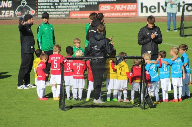 Zwei Gruppen von Kindern in Fu&szlig;balltrikots stehen nebeneinander auf einem Rasenplatz, umgeben von Trainern.