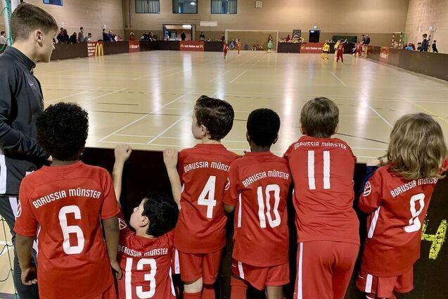 Sechs junge Fu&szlig;ballspieler in roten Trikots mit der Aufschrift &bdquo;Borussia M&uuml;nster&ldquo; beobachten das Spiel von der Seitenlinie.
