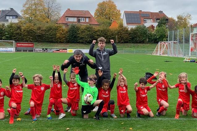 Gruppe von Kindern in roten Fu&szlig;balltrikots posiert jubelnd auf einem Rasenfeld mit Toren im Hintergrund.
