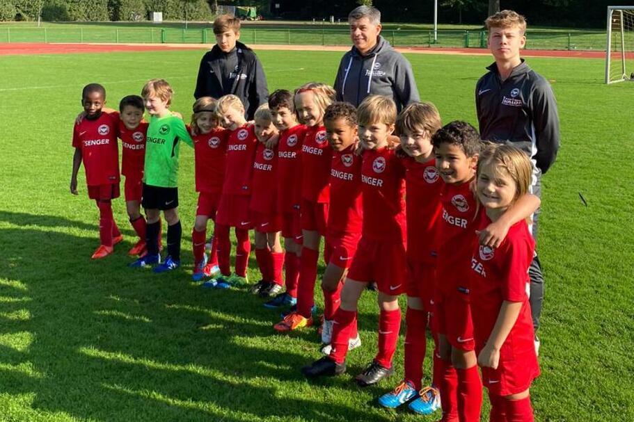 Gruppe von Kindern in roten Fu&szlig;balltrikots mit Trainern auf einem Sportplatz, umgeben von B&auml;umen und einem Tor.