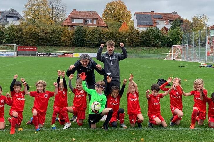 Gruppe von Kindern in roten Fu&szlig;balltrikots posiert jubelnd auf einem Rasenfeld mit Toren im Hintergrund.