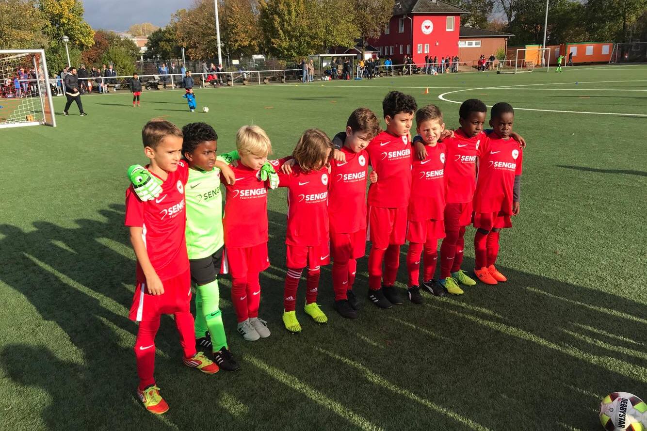 Gruppe von acht jungen Fu&szlig;ballspielern in roten Trikots, posierend auf einem Spielplatz mit Rasen.