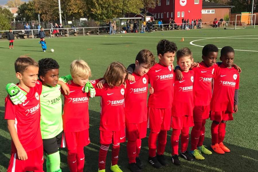Gruppe von acht jungen Fu&szlig;ballspielern in roten Trikots, posierend auf einem Spielplatz mit Rasen.