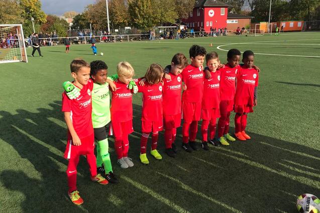 Gruppe von acht jungen Fu&szlig;ballspielern in roten Trikots, posierend auf einem Spielplatz mit Rasen.