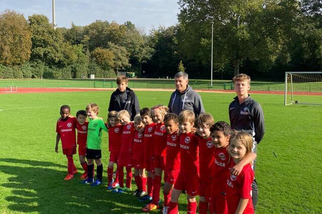 Gruppe von Kindern in roten Fu&szlig;balltrikots mit Trainern auf einem Sportplatz, umgeben von B&auml;umen und einem Tor.