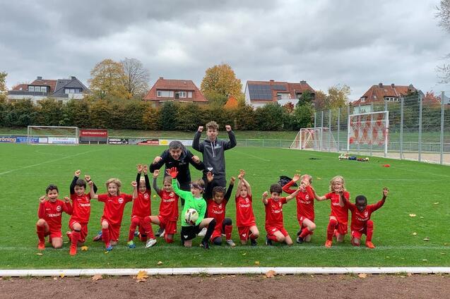 Gruppe von Kindern in roten Fu&szlig;balltrikots posiert jubelnd auf einem Rasenfeld mit Toren im Hintergrund.